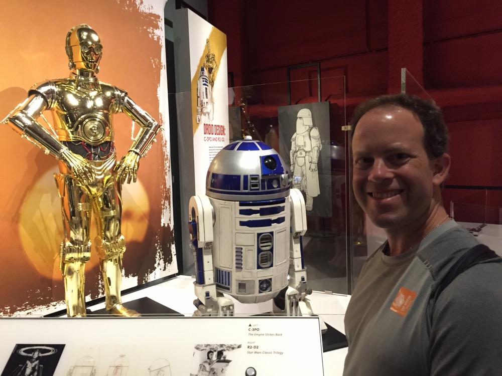 Russ Rosenzweig standing beside the C-3PO and R2-D2 props from the Star Wars films at a museum exhibit