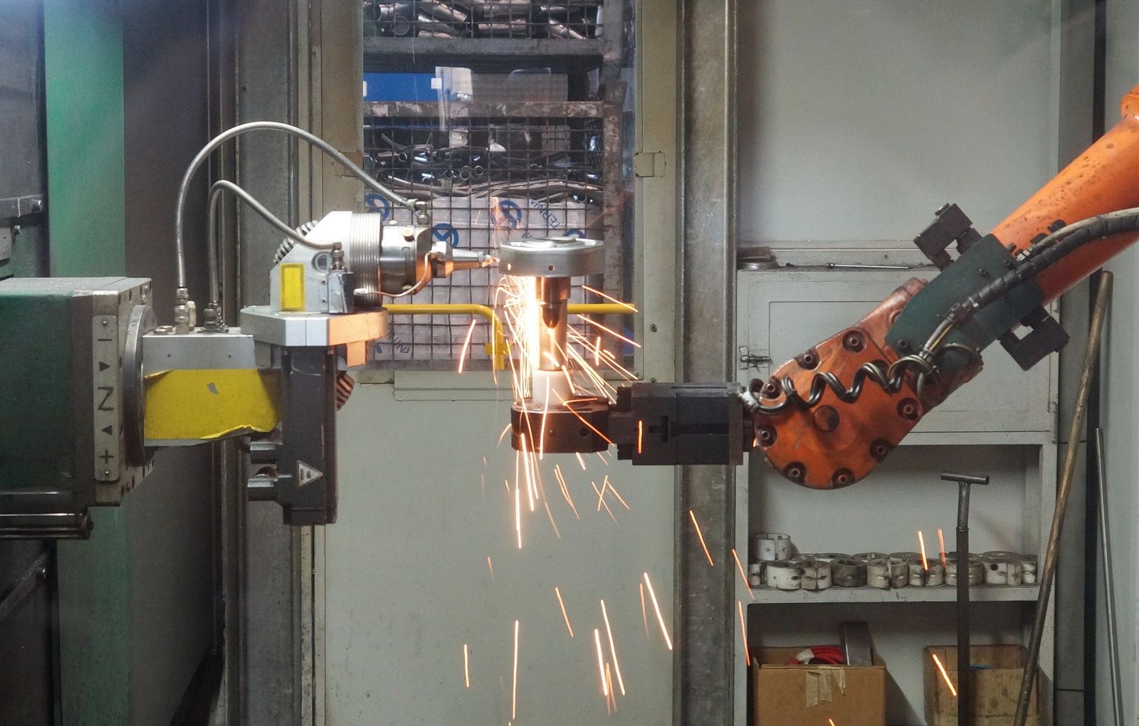 A robotic welding cell at work, sparks flying as an orange industrial arm holds a part against a spindle